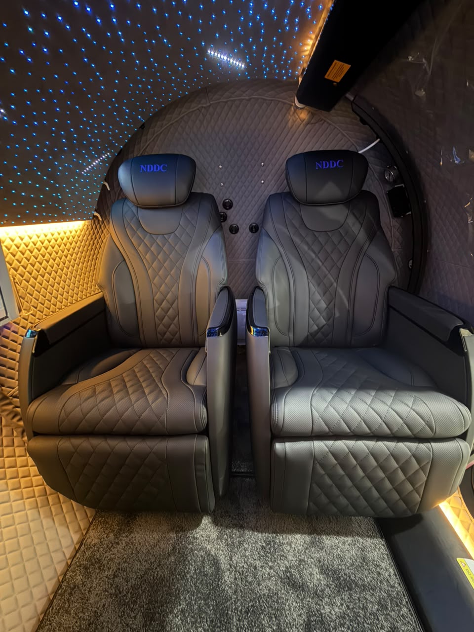 Head-on view of two dark quilted-leather reclining captain's chairs with embroidered NDDC headrests and a center console, set beneath a starlight ceiling with gold-padded interior lining