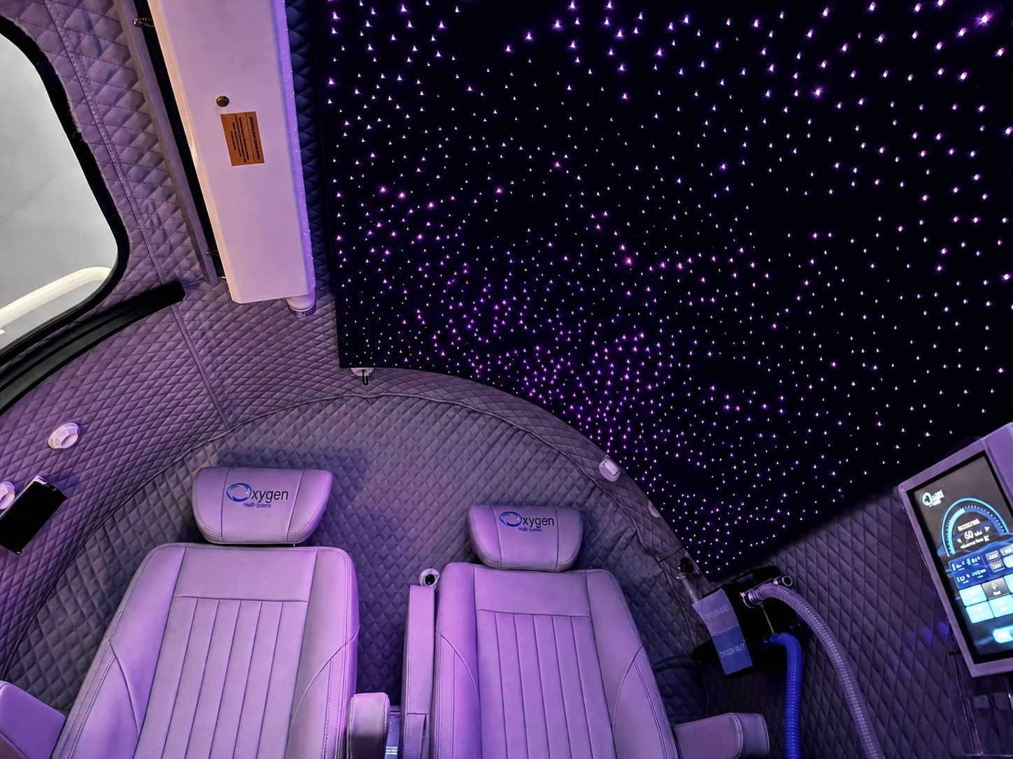 Interior view of the M5502 chamber's fiber-optic starlight ceiling glowing with purple star points above two lavender quilted-leather seats with embroidered Oxygen Health Systems headrests and the touchscreen control panel