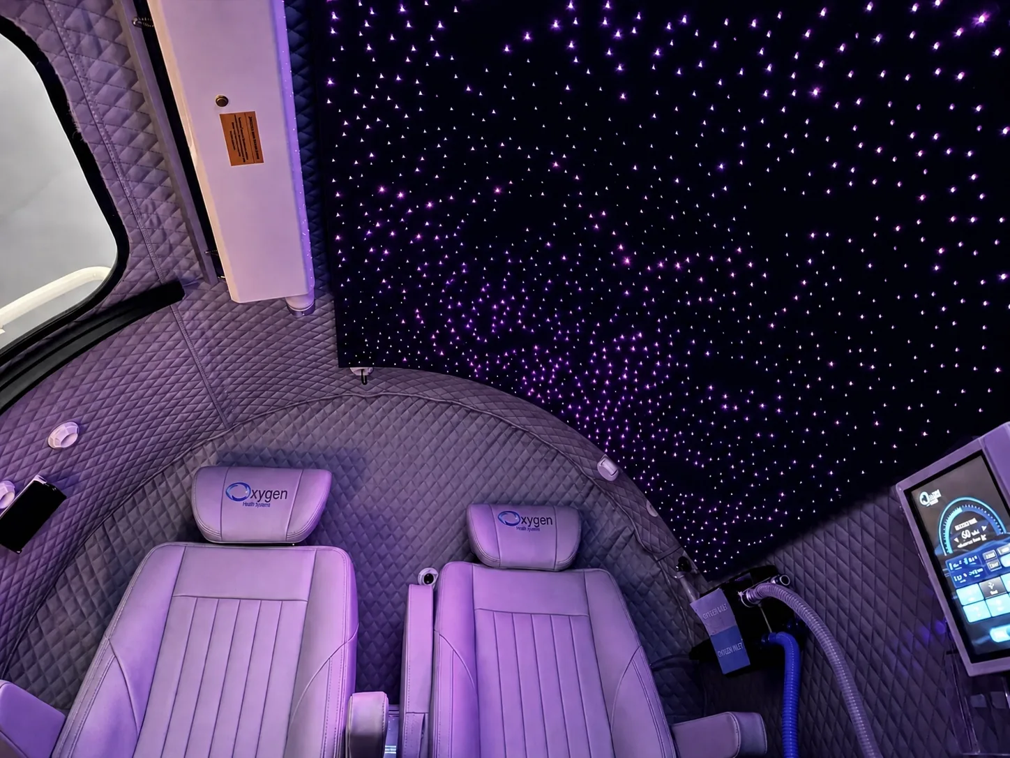 Interior view of the M5502 chamber's fiber-optic starlight ceiling glowing with purple star points above two lavender quilted-leather seats with embroidered Oxygen Health Systems headrests and the touchscreen control panel